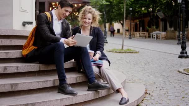 Usmívající se velké dobře vypadající dáma a kluk studenti se posadit na schodech uprostřed ulice a pomocí elektronického tabletu analyzovat něco — Stock video