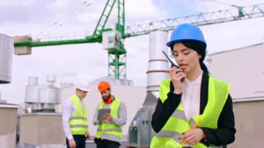 Modern inşaat alanındaki karizmatik kadın bir radyo telekomünikasyonundan bazı inşaat işleriyle uğraşırken arka plandaki diğer uzmanlar diğer plan detaylarını tartışıyor.