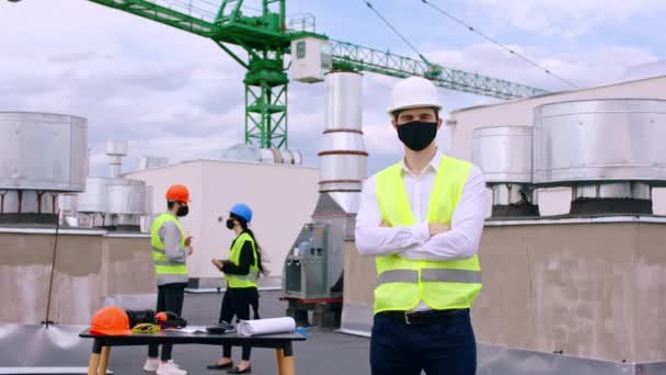 屋根の上の建設現場でのCovid-19のコンセプトカメラの前にポーズをとる主な建築家他の専門家顔に保護マスクで計画のいくつかの詳細を議論 — ストック動画