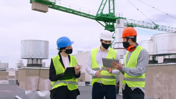 Группа инженеров-технологов COVID-19, анализирующих план строительства, обсуждающих некоторые детали с использованием цифровых планшетов в защитных шлемах — стоковое видео