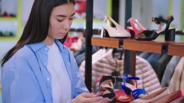 Attractive young woman asian looking in a shoe shop take some pair of shoes from the shelves concentrated she have a shopping day — Stock Video