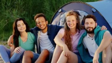 Büyük bir arkadaş grubu piknikte kameranın önünde poz veriyor. Büyük bir çadırın yanında oturuyorlar.