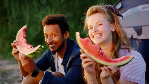Tipul afro-american și prietena lui drăguță se bucură de timpul petrecut împreună la picnic stau pe iarbă uitându-se direct la cameră și zâmbind mare în timp ce țin câteva bucăți gustoase de — Videoclip de stoc
