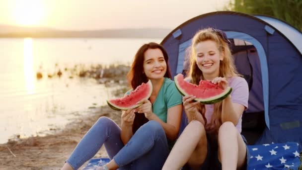 Ładne panie przed kamerą na pikniku obok niesamowitego jeziora i zachodu słońca trzymając duże kawałki arbuza i uśmiechając się duże — Wideo stockowe