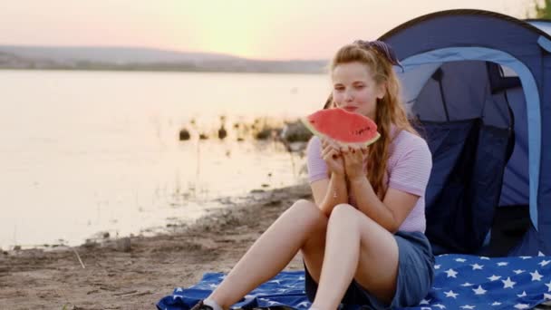 Charismatic lady beside the lake at sunset take a sit down beside the tent at the picnic she holding a big piece of watermelon and looking straight to the camera. Shot on ARRI Alexa — Stock Video