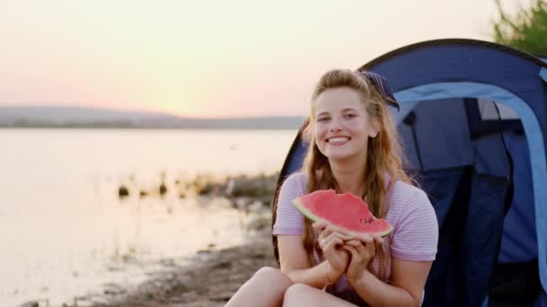 Piękna blondynka z dużym uśmiechem trzymająca duży kawałek arbuza siedząc obok namiotu o zachodzie słońca — Wideo stockowe