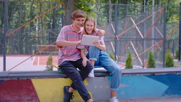 Charismatic casal bonito em um parque de skate moderno sentado nas cadeiras passar o tempo juntos fora eles usando um gadget tablet eletrônico — Vídeo de Stock