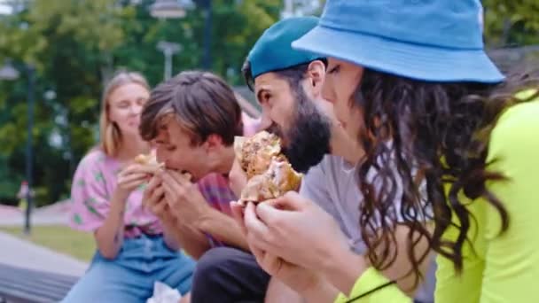 कैमरा के सामने क्लोजअप एक शहरी स्केट पार्क में हैम्बर्गर खाने वाले दोस्तों के युवा समूह को स्केटबोर्डिंग शुरू करने से पहले उनके पास ब्रेक है — स्टॉक वीडियो