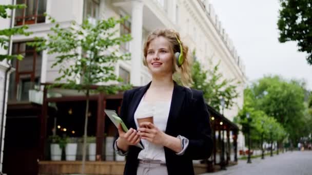 Em uma rua moderna jovem mulher andando até a rua ela segurando o tablet eletrônico e café sorrindo bonito — Vídeo de Stock