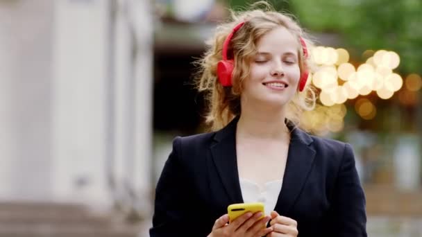 Hermosa joven delante de la cámara en medio de la calle bailando y escuchando música desde unos grandes auriculares rosados — Vídeo de stock