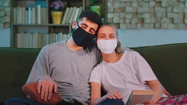Casal atraente em casa em pandemia com uma máscara protetora olhando diretamente para a câmera enquanto sentado no conceito de sofá de Coronavirus — Vídeo de Stock