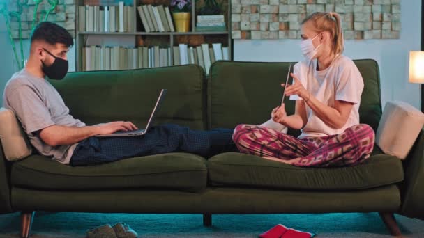 Pandemisches Coronavirus-Paar bleibt zu Hause auf dem Sofa, auf dem sie arbeiten und gleichzeitig online studieren, mit Laptop und Tablet und Schutzmaske — Stockvideo