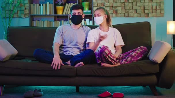 En casa en la pareja pandémica viendo la televisión sentada en el sofá que llevaba el concepto de máscara protectora de Covid-19 — Vídeos de Stock