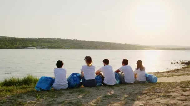 晴れた日のボランティアのグループで素晴らしい湖の景色で彼らはビーチに座って湖を見て休憩を取ります — ストック動画