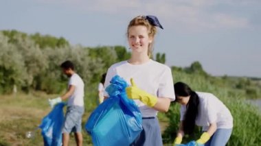 Kameraya yakın çekim yapan genç gönüllü kadın kameranın önünde büyük mavi bir çanta tutuyor ve arkadaşları gönüllü olarak mavi plastik poşetleri alıyor ve topluyor.