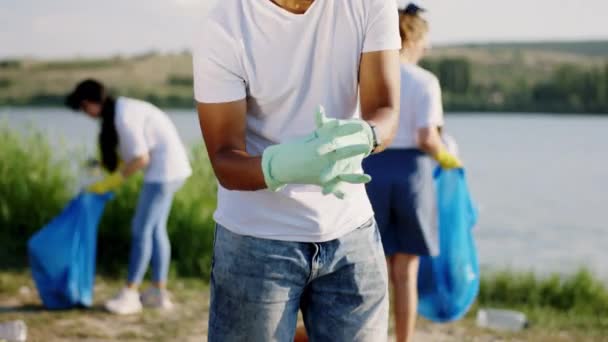 Detaljer om en volontär kille afro American bär skyddshandskar och ta de stora blå plastpåsar börjar plocka upp skräp från stranden sidan — Stockvideo