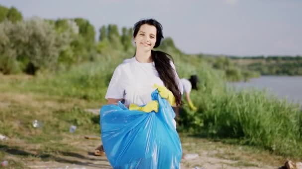 Hermosa señora voluntaria con una gran bolsa de plástico azul recogiendo la basura de la playa junto al lago con sus diversos amigos voluntarios que limpian la naturaleza de la basura — Vídeo de stock