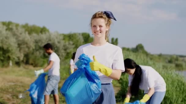 Κοντινό πλάνο στην κάμερα νεαρή εθελόντρια κρατώντας μια μεγάλη μπλε τσάντα μπροστά από την κάμερα και οι φίλοι της εθελοντές μαζεύοντας την αρπαγή και τη συλλογή από τις μπλε πλαστικές σακούλες — Αρχείο Βίντεο