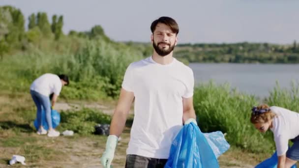 Postarejte se o naši planetu skupina dobrovolníků chlap dívá přímo do kamery, zatímco sbírat odpadky drží velký modrý plastový sáček a nosí plastové rukavice — Stock video