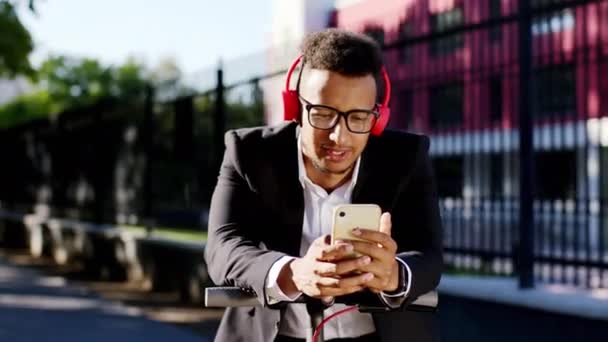 Retrato de um estudante afro-americano ouvindo música usando grandes fones de ouvido vermelhos ele aproveitando o tempo em sua scooter elétrica no meio da rua — Vídeo de Stock