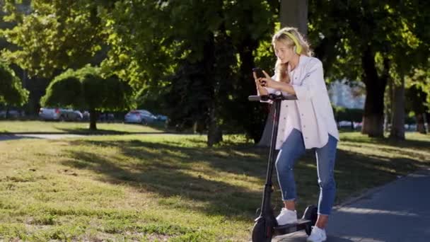 W słoneczny dzień na środku ulicy niesamowicie wyglądająca blondynka zatrzymuje się na skuterze elektrycznym, rozmawia na swoim smartfonie wpisując wiadomość. Strzał na ARRI Alexa Mini — Wideo stockowe