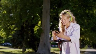 Modern bir caddenin ortasında güneşli bir günde güzel bir kadın akıllı telefonunu kullanarak bir mesaj yazıyor. Çok mutlu ve büyük bir gülümsemeyle elektrikli scooterına biniyor.