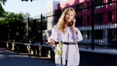 Güzel, gülümseyen, sarışın, güzel bir kız öğrenci telefonda konuşmak için elektrikli scooterını durdurdu. Gülümsedi ve çok heyecanlandı. ARRI Alexa Mini 'de çekilen