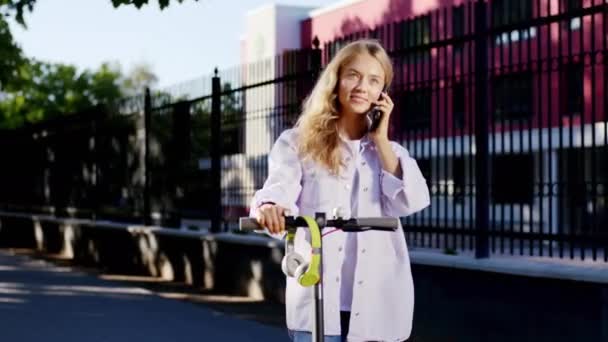 Atraktivní s krásným úsměvem blond vlasy studentka udělat zastávku na její elektrický skútr, aby konverzaci na telefonu se usmála velké a pocit, že velmi vzrušený. Shot na ARRI Alexa Mini — Stock video