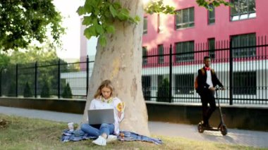 Caddenin dışında Afro-Amerikalı bir ofis çalışanı elinde elektrikli scooterla çimlere oturmuş bilgisayarını çalıştıran bir kadınla iş arkadaşına geldi.