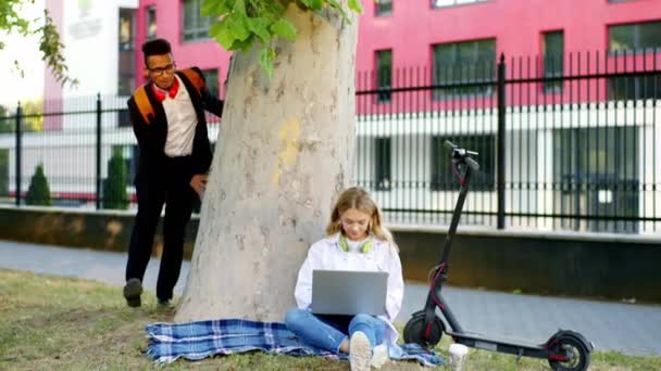 Zabawne studenci koledzy Afro amerykański facet zrobić kopa do jego kolega ładna pani na zewnątrz obok uczelni podczas gdy pani wpisując coś na laptopie dół na trawie — Wideo stockowe