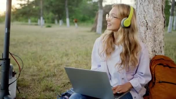 Σε εκπληκτικό μέρος στη φύση ελκυστική ξανθιά μαθήτρια μαλλιά κάνουν ένα διάλειμμα σε καθαρό αέρα ακούγοντας μουσική από ακουστικά και πληκτρολογώντας κάτι στο φορητό υπολογιστή. Πυροβολήθηκε στην ΑRI Αλέξα — Αρχείο Βίντεο