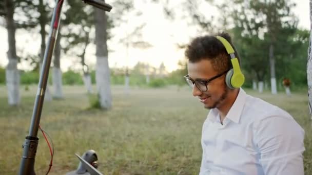 Charismatique avec un grand sourire Afro-Américain avec un gros casque faire une pause sur le parc après le cours universitaire profiter du temps à la nature, il est venu avec le scooter électrique. 4k — Video