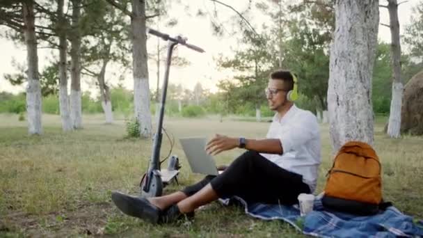 En kille bra efter arbetet kom i parken med elektrisk skoter ta en sitta ner på gräset med hjälp av den bärbara datorn för att göra en videochatt han visar något med hjälp av webbkameran han bär stora — Stockvideo