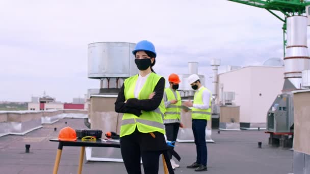 Pandemia di COVID-19 bella donna ingegnere in cantiere sulla parte superiore dell'edificio in piedi di fronte alla telecamera con maschera protettiva e caschi di sicurezza. Girato su ARRI Alexa — Video Stock