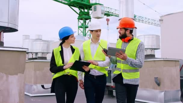 Moderno canteiro de obras no topo da construção de jovens especialistas andando pelo local analisando usando um tablet dígito alguns detalhes da construção que eles usam capacetes de segurança. Tiro em ARRI — Vídeo de Stock