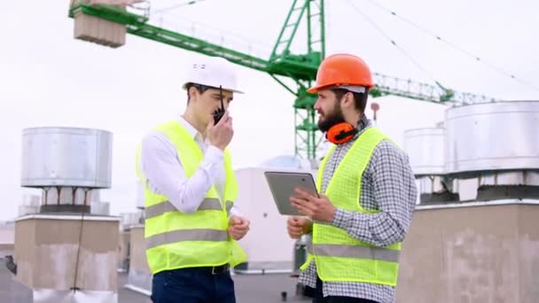 Good looking guys young specialists of construction site on the roof top while analyzing the plan of construction using digital tablet start walking through the site and using a radio for — Stock Video