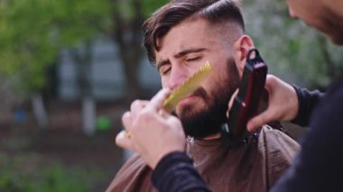 Kameranın önünde berber bahçedeki adama sakal yapıyor. Elektronik makas kullanıyor.