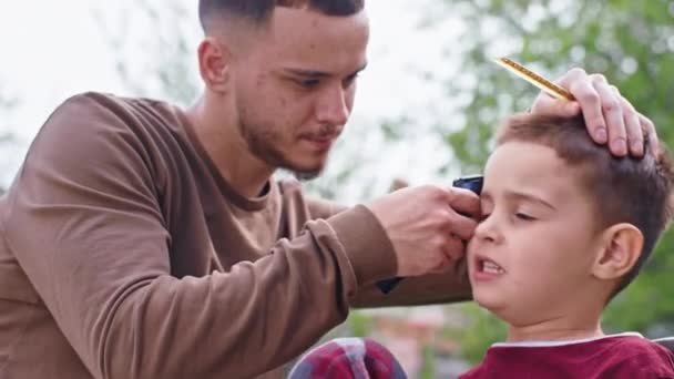 Перед камерой смешной маленький мальчик сделать стрижку в саду он сидит на стуле и парикмахер с клиппером закончить стрижку волос — стоковое видео