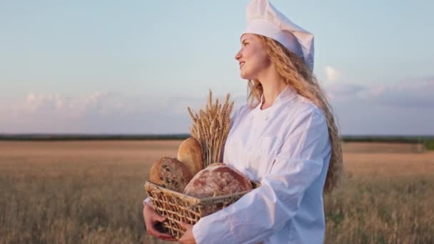 Close-up charismatisch met een mooie glimlach dame bakker met krullend lang haar met een mand vol vers gebakken brood in het midden van tarweveld — Stockvideo