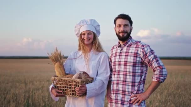 Портрет мужчины-фермера и пекаря перед камерой, стоящей вместе леди, держащей корзину со свежим хлебом посреди пшеничного поля — стоковое видео