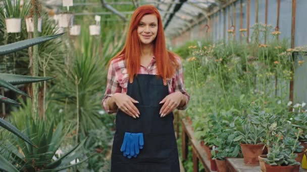 Krásná zahradnice ve stylové uniformě stojící uprostřed skleníku květin a dívající se přímo do kamery pózující — Stock video