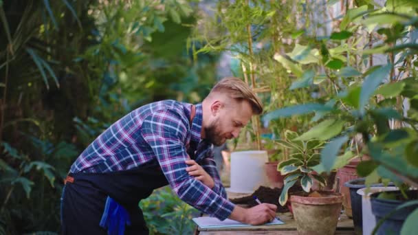Großartig aussehender Gärtner im eigenen Gewächshaus macht sich Notizen über den Zustand der Dekoration, die er auf der Karte aufschreibt — Stockvideo