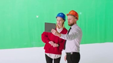 Yeşil arka plan stüdyosu güzel bayan asistan ve güvenlik kasklı adam mimar inşaat planını analiz etmek için dijital tablet kullanıyor.