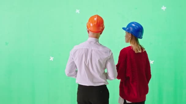 漂亮的男建筑师和他的助理女士在一个彩色钥匙工作室里分析他们戴着安全帽的工地的建设计划 — 图库视频影像