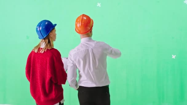Jonge zakenman en een dame ingenieur in een groene achtergrond studio met een veiligheidshelmen analyseren van het plan van de bouwplaats met een digitale tablet. Neergeschoten op ARRI Alexa — Stockvideo