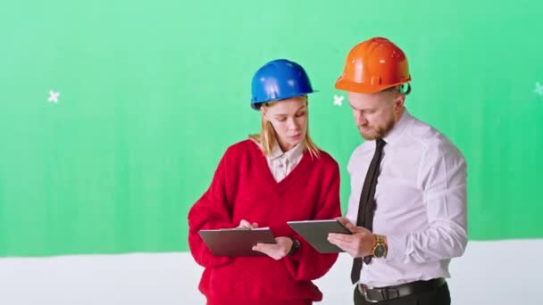 녹색 배경 스튜디오에 있는 두 명의 엔지니어 숙녀와 남성 디지털 태블릿을 사용하여 안전 헬멧을 쓰고 건설 현장의 계획을 분석하는 — 비디오