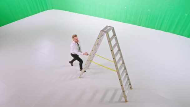 In een grote groene studio zakenman met een grote glimlach klimt hij een ladder terwijl hij recht naar de camera kijkt — Stockvideo