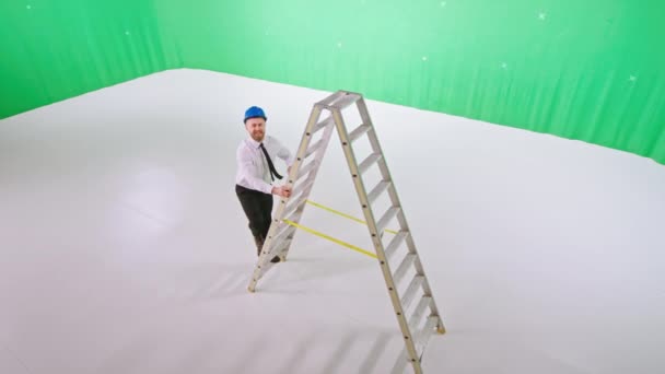 Knappe architect met een veiligheidshelmen poserend voor de camera terwijl hij een ladder beklimt in een grote groene achtergrond studio — Stockvideo