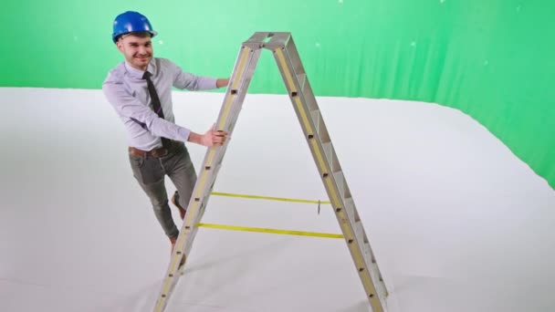 Усміхнений хлопець інженер позує перед камерою, стоячи на драбині у великій зеленій фоновій студії — стокове відео