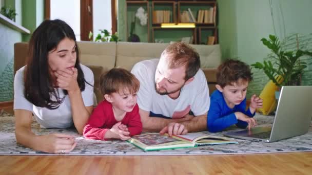 Перед камерою вниз на підлозі у вітальні мама і тато готують історію з книги до своєї маленької дитини і в той час як інша велика дитина використовує ноутбук, щоб грати в гру — стокове відео